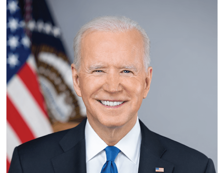 President Joe Biden wearing a dark blue suit and light blue tie, with a white shirt and a flag pin. He is standing in front of a gray background with two flags. On the left the U.S. flag and on the right the presidential seal flag