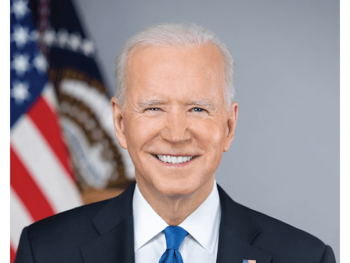 President Joe Biden wearing a dark blue suit and light blue tie, with a white shirt and a flag pin. He is standing in front of a gray background with two flags. On the left the U.S. flag and on the right the presidential seal flag